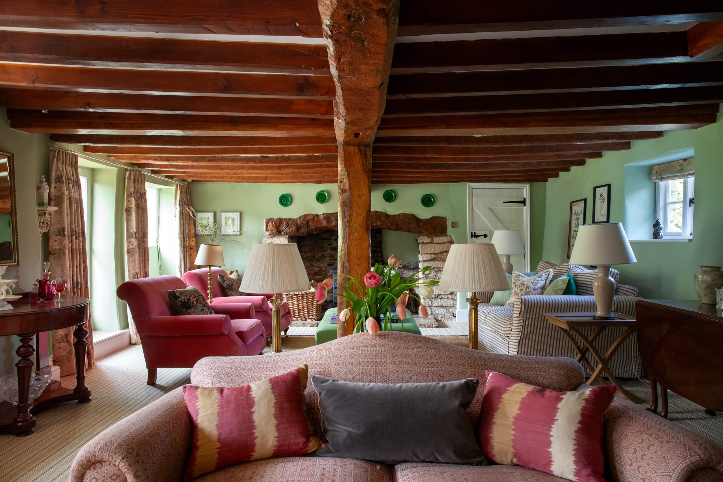 A cozy cottage living room by Phoebe Bray Interiors features exposed wooden beams, a stone fireplace with a wood-burning stove, green walls with decorative plates, comfortable seating, a green ottoman, fresh flowers, and books.