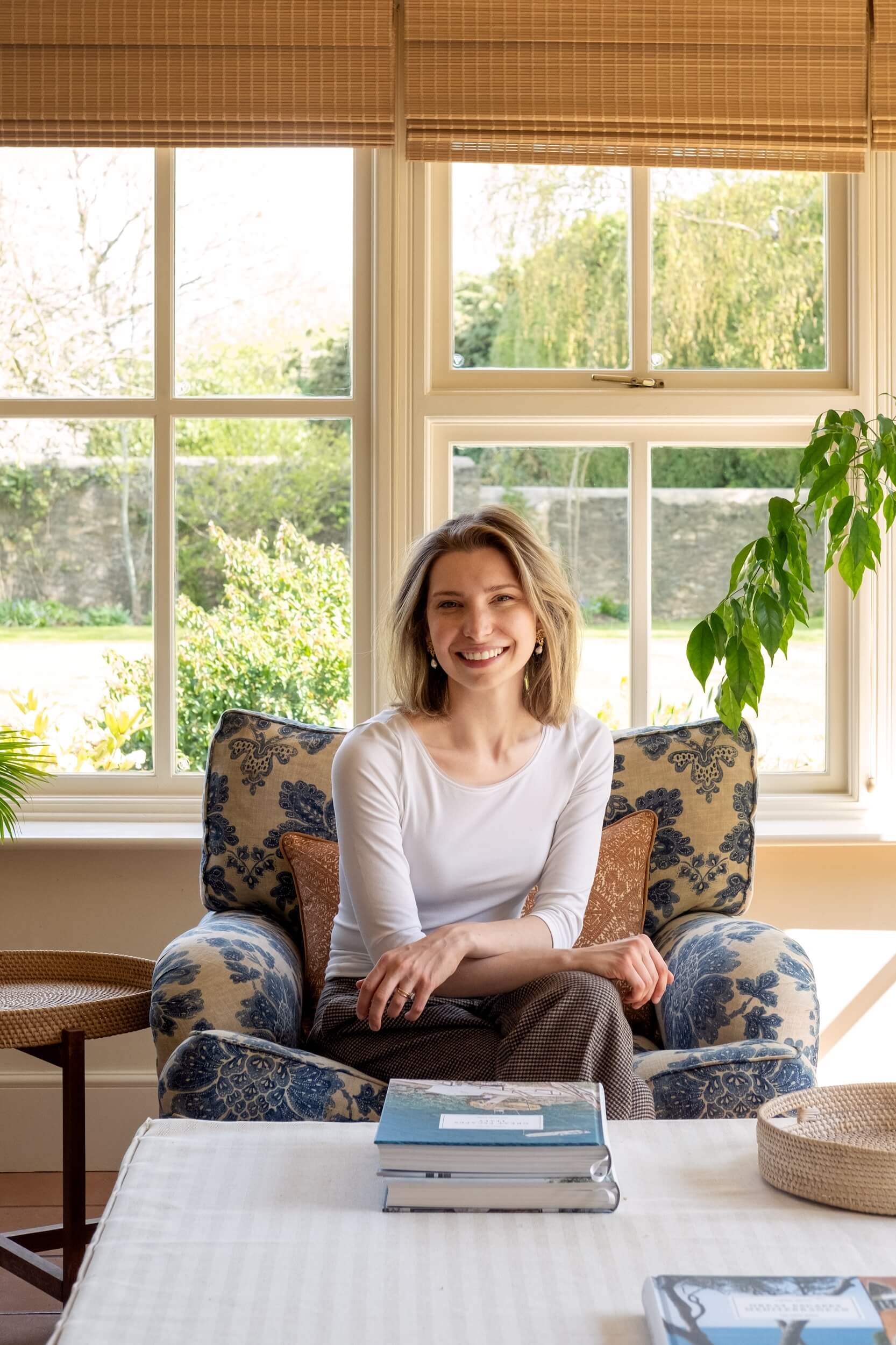 A woman with blonde hair, Phoebe Bray, wearing a white long-sleeved shirt and brown trousers, sits smiling on a blue patterned armchair in a bright room designed by Phoebe Bray Interiors, with large windows and lush greenery outside.